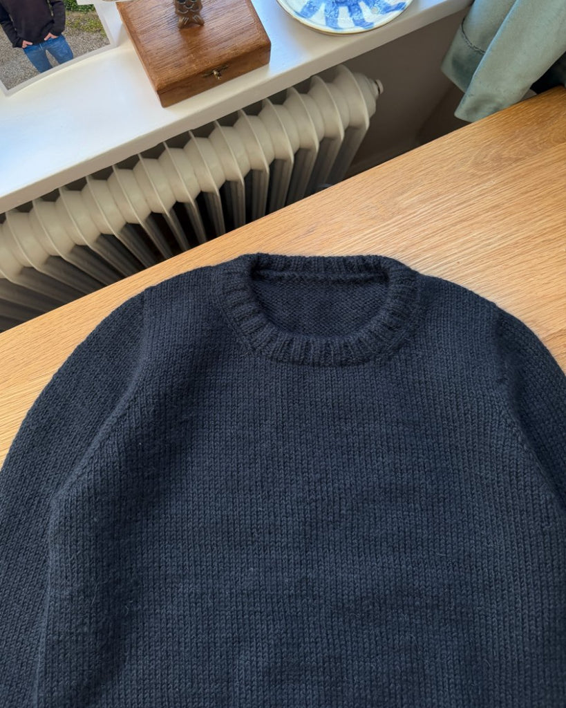 Blå Niels Sweater Junior liggende på skrivebord med fokus på halskanten